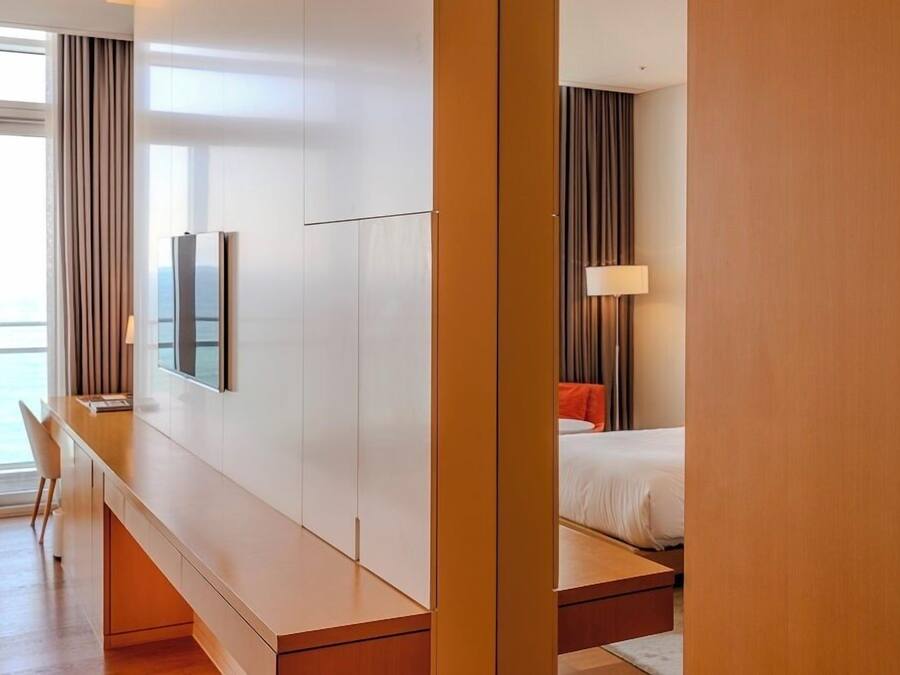 A modern hotel room with a wooden desk, a bed with a red pillow, and a view of the sea through large windows.