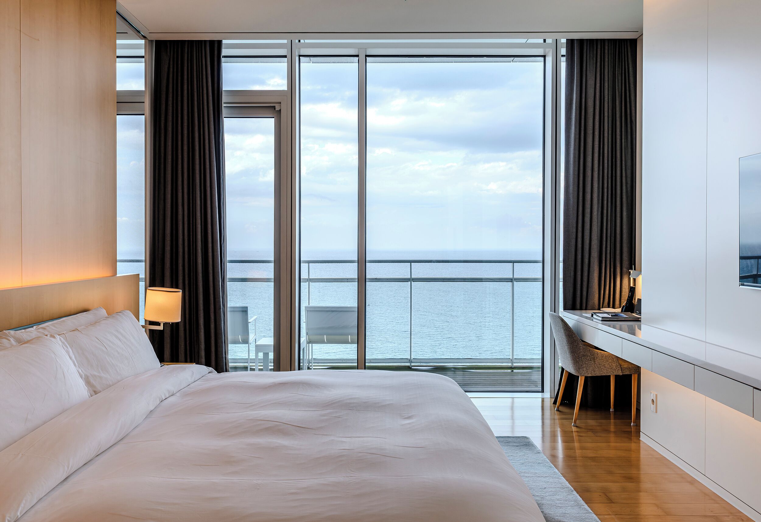 A hotel room with a large bed, a desk with a chair, a TV, and a view of the sea.