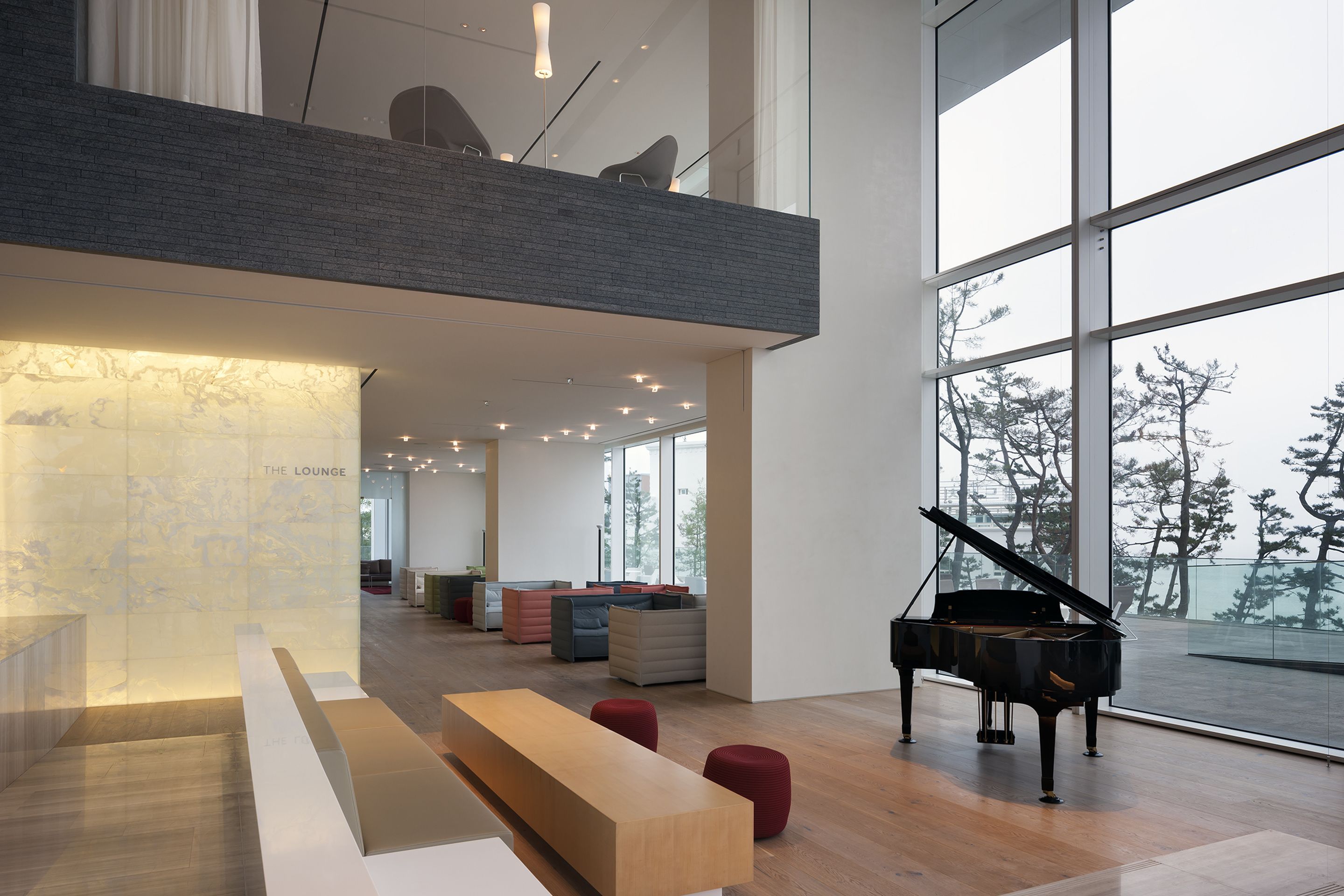 Eine großzügige Lounge mit einem Flügel, Holzbänken und einer hohen Decke mit großen Fenstern, die einen Blick auf Bäume gewähren.