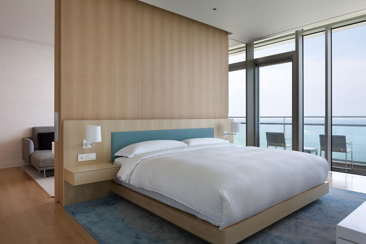 Quarto de hotel moderno com uma cama grande, uma mesinha de cabeceira, uma cadeira e vista para o mar através de amplas janelas.