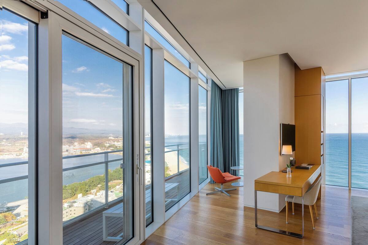 A modern hotel room with large floor-to-ceiling windows, a wooden desk, a chair, and a view of the sea.