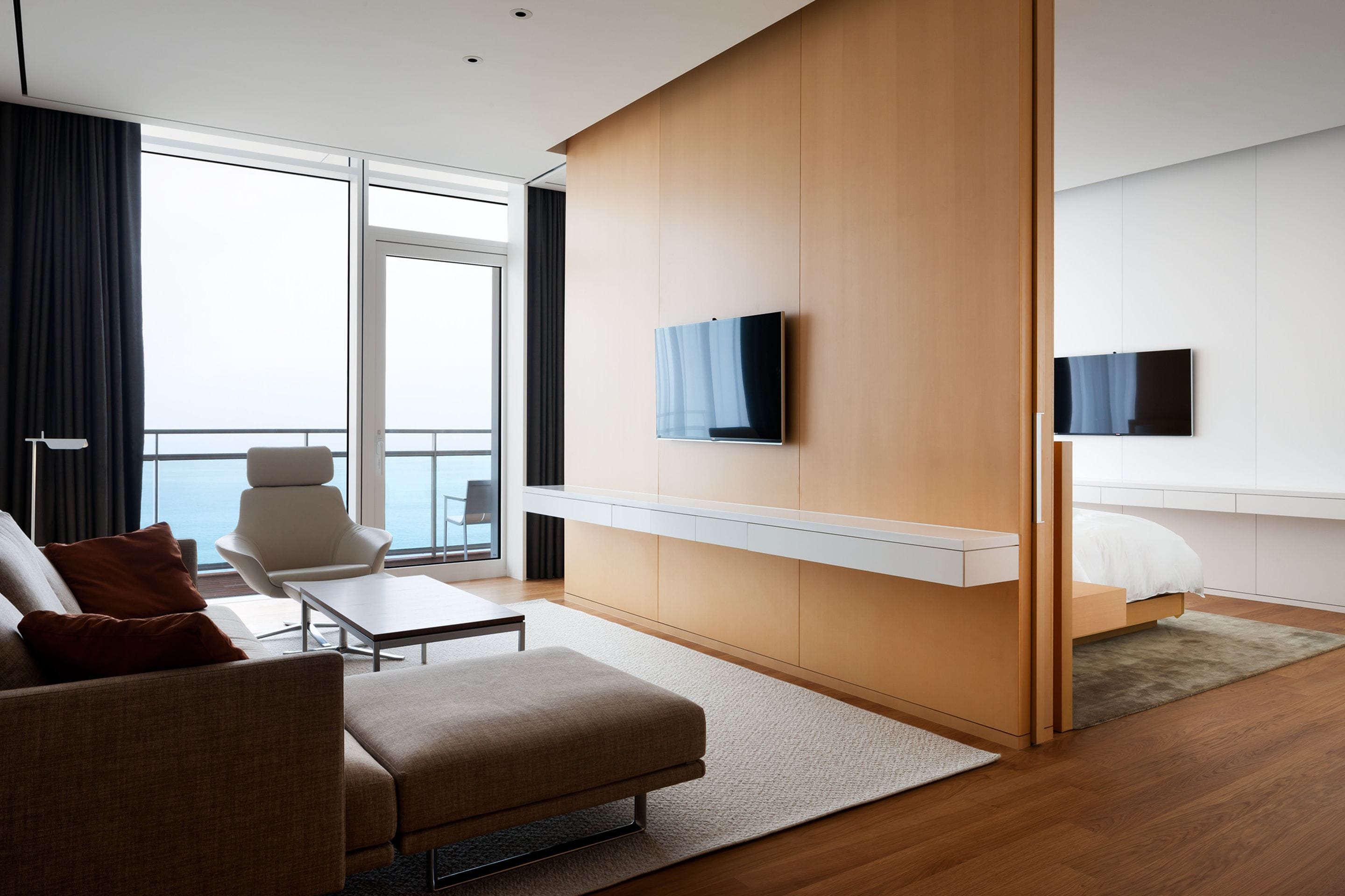 A modern hotel room with a sofa, a small table, a TV mounted on the wall, and a bed with a white headboard.