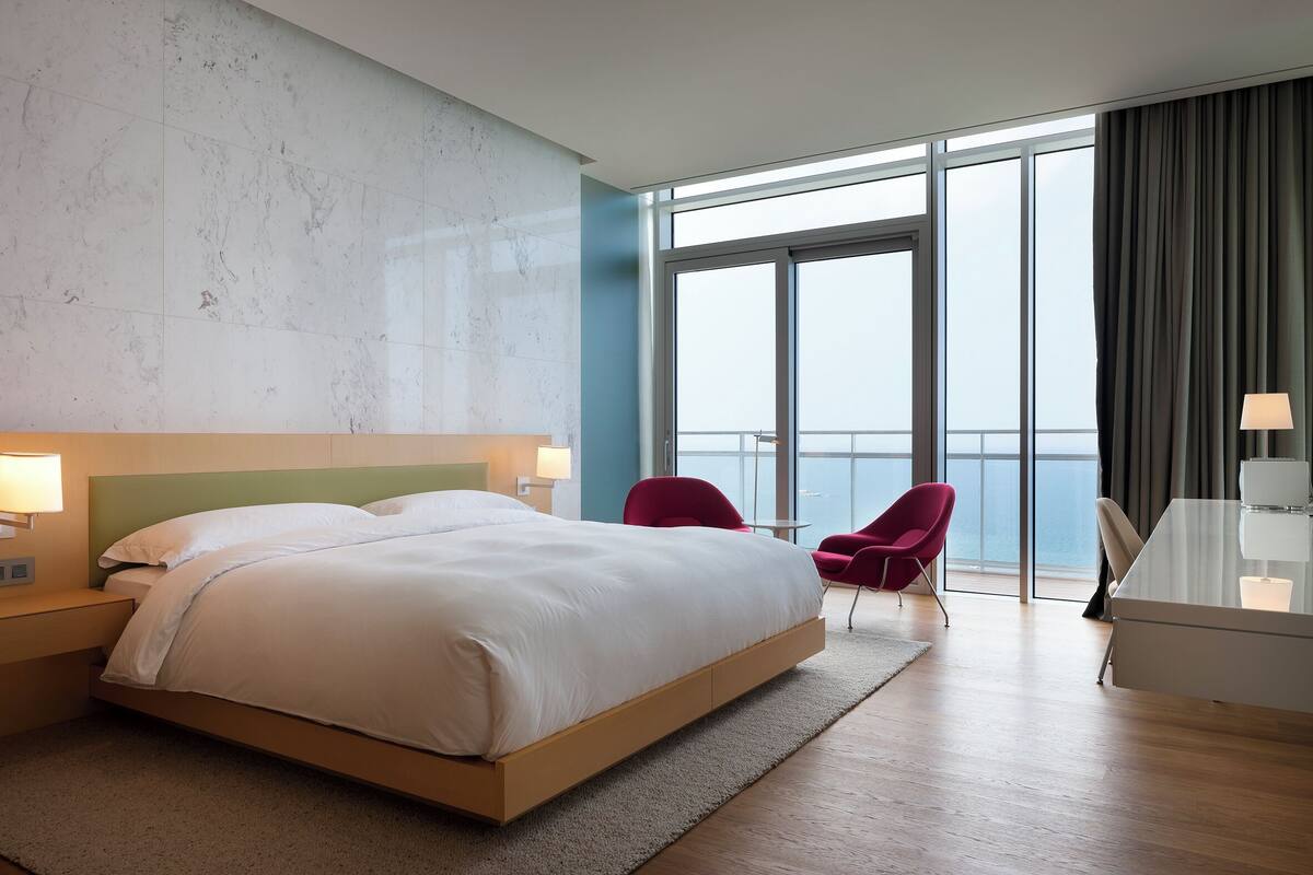 Quarto de hotel moderno com uma cama grande, uma escrivaninha e vista para o mar por grandes janelas.