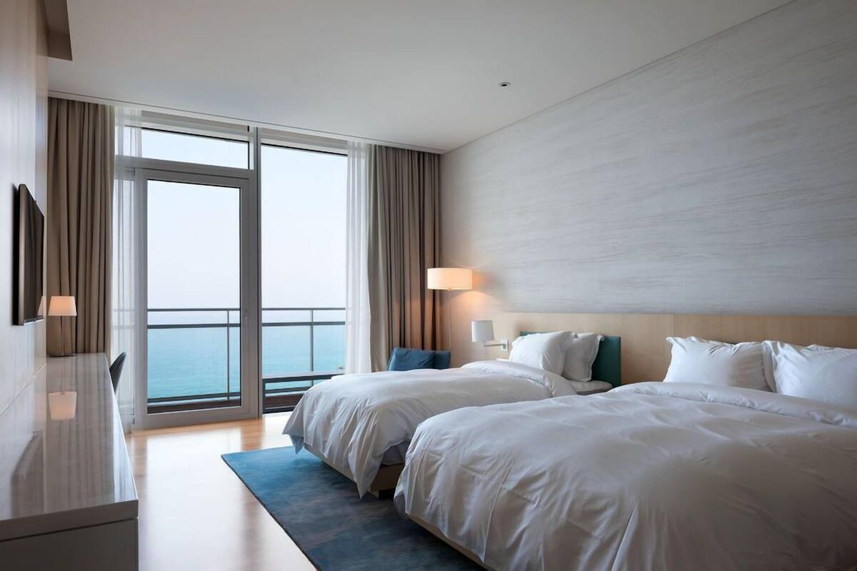 Quarto de hotel com duas camas, uma escrivaninha, uma luminária e uma janela grande com vista.