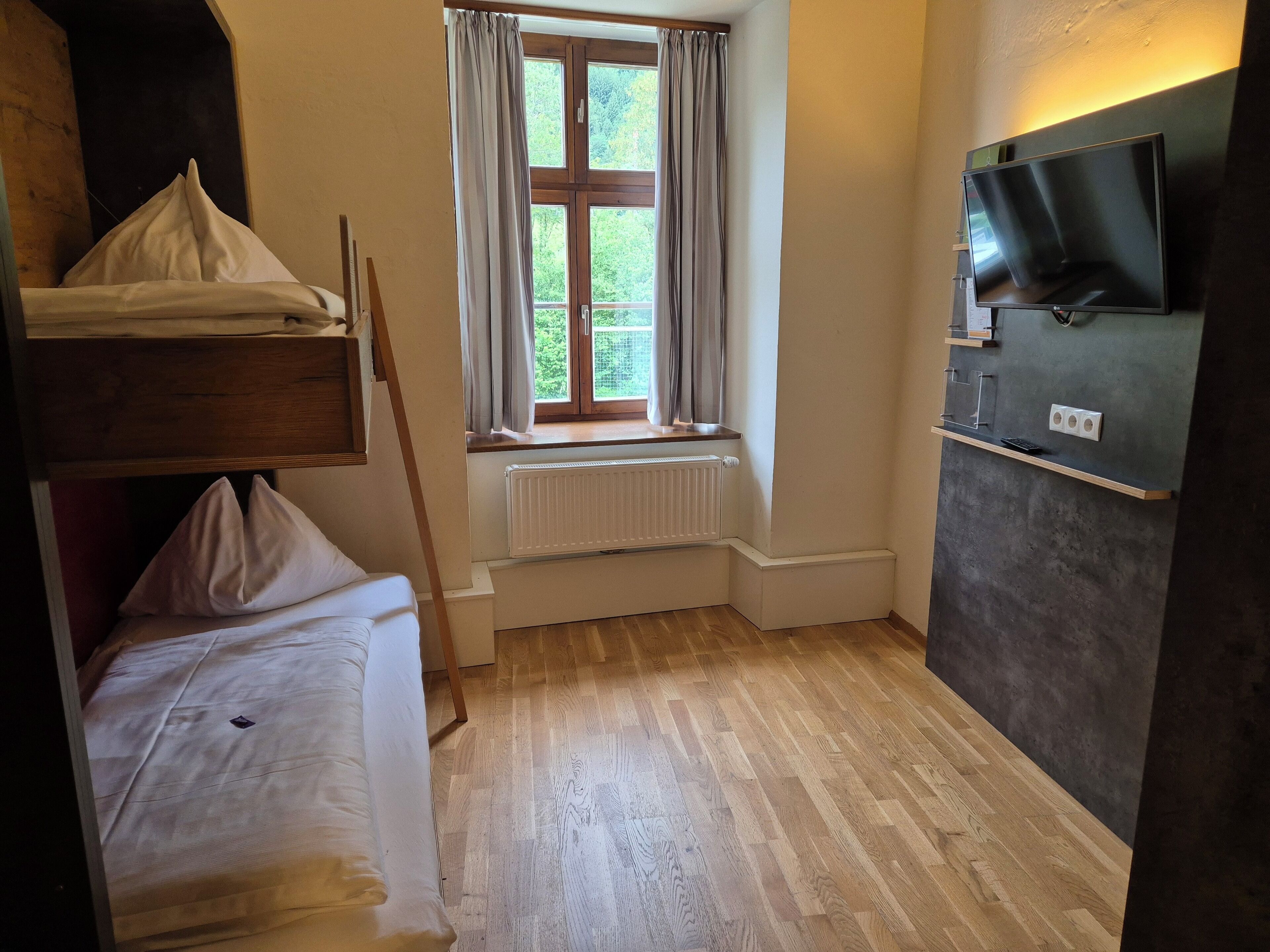 A small room with a bunk bed, a window with curtains, a radiator, a television mounted on the wall, and a wooden floor.