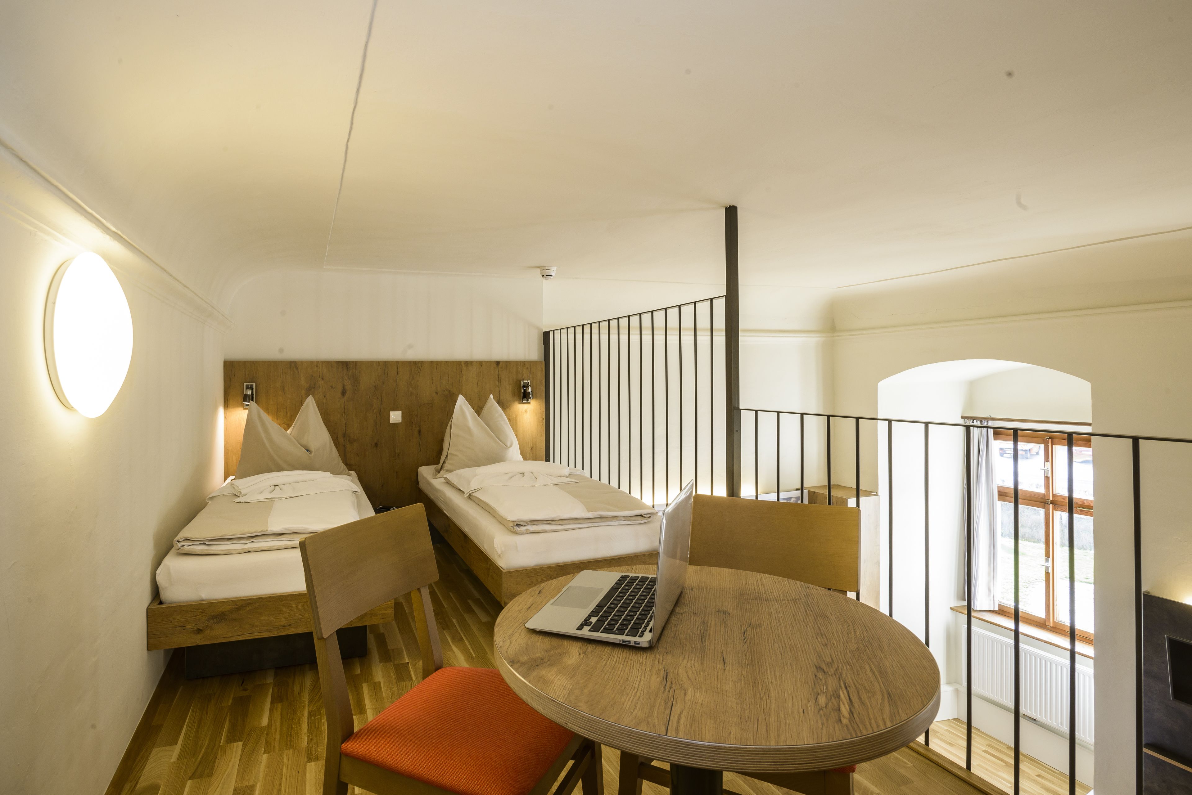 Een kleine kamer met twee eenpersoonsbedden, een ronde houten tafel met een laptop, en uitzicht op het omliggende landschap door een raam.
