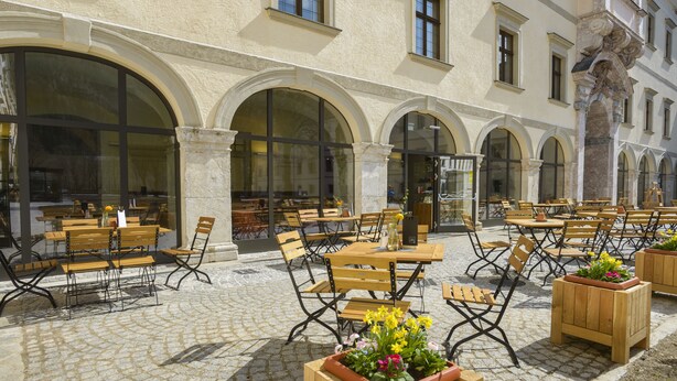 An outdoor cafe with wooden tables and chairs, large arched windows, and potted plants.