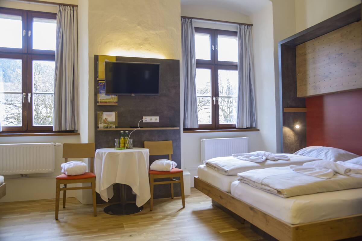Una habitación con una cama de madera, una mesa con una silla, un televisor montado en la pared y ventanas con cortinas.