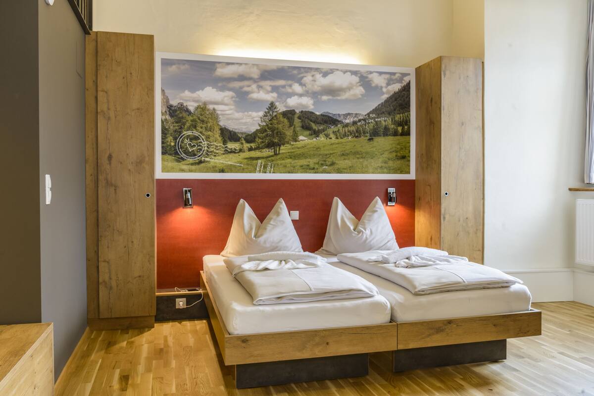 Una cama doble con ropa blanca, una pared de acento roja y un cuadro de paisaje grande encima.