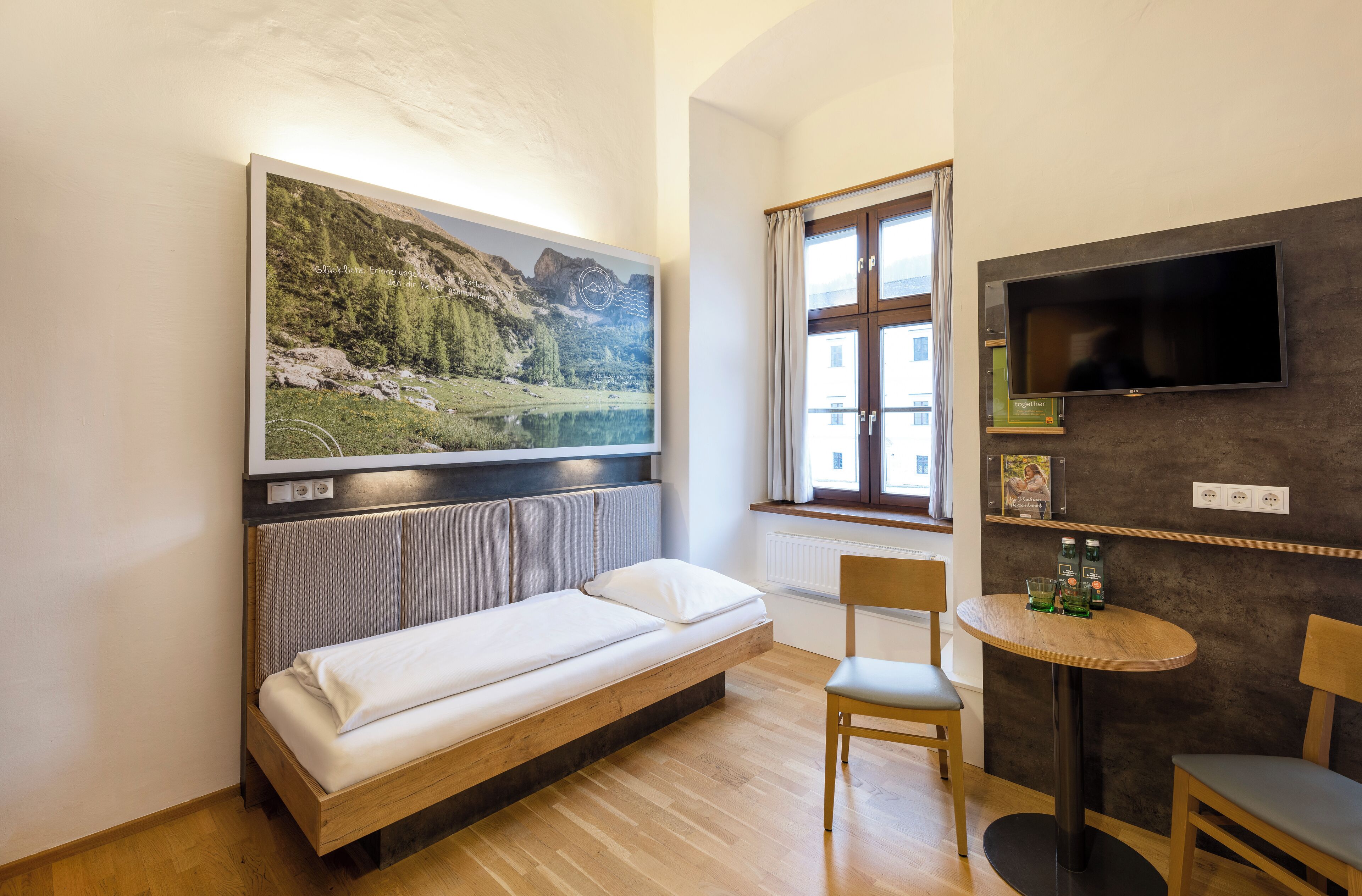 A modern room with a bed, a TV mounted on the wall, a window with curtains, and a small table with chairs.