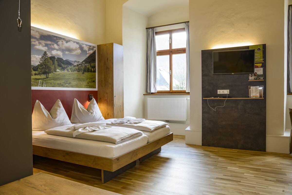 Un dormitorio con una cama de madera, un cuadro de paisaje grande, una ventana con cortinas, un televisor y un radiador.