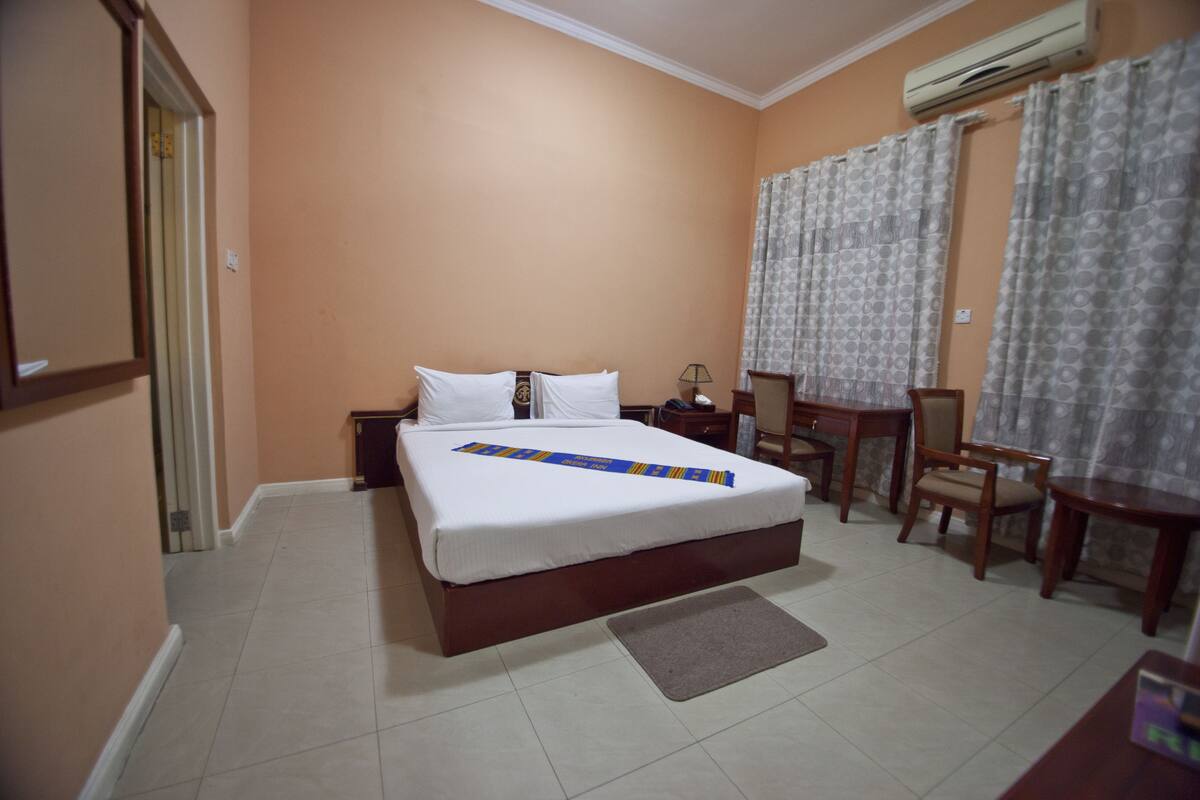 A hotel room with a bed, a desk, a chair, and an air conditioning unit.
