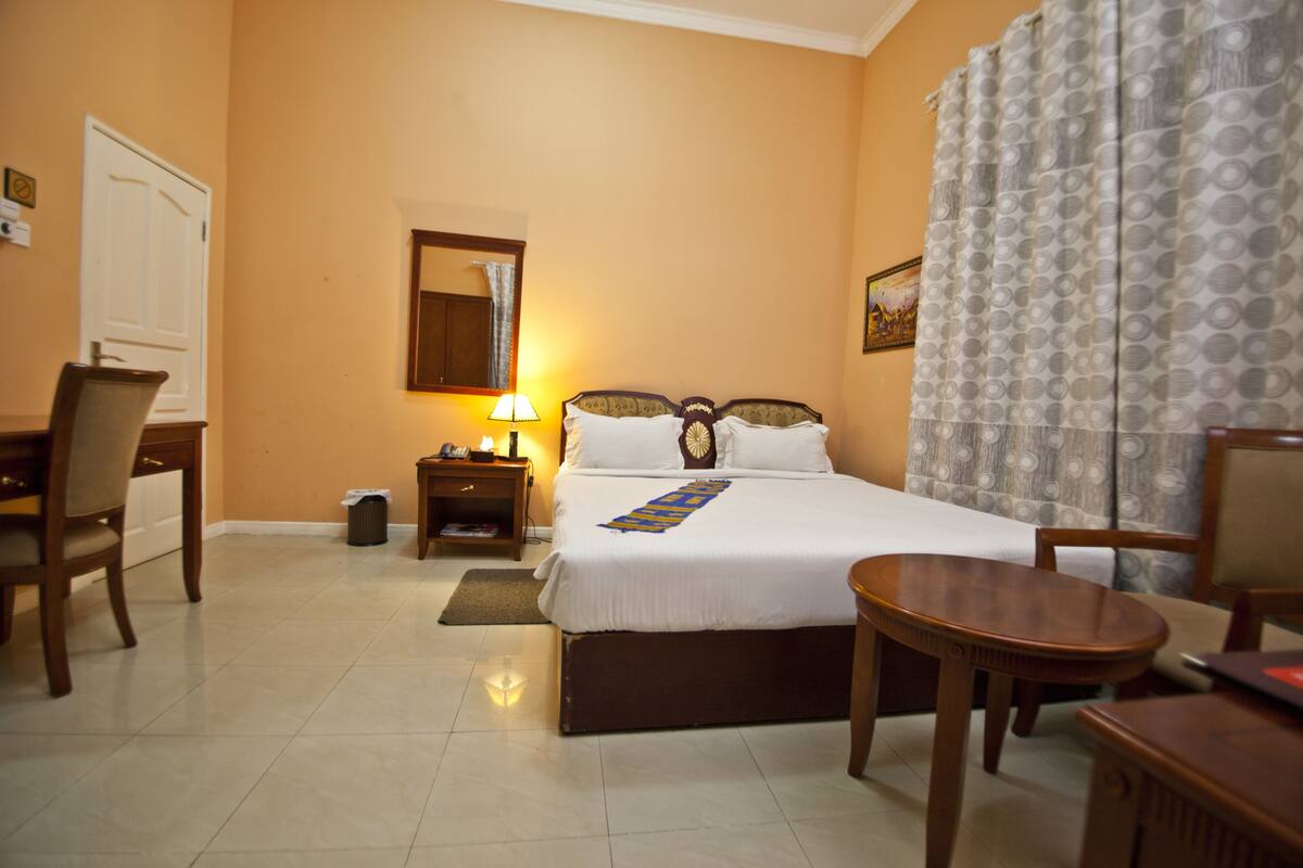 A hotel room with a bed, a desk, a chair, a nightstand, and a lamp.