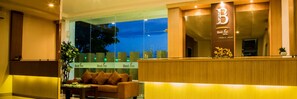 A hotel reception area with a wooden reception desk, a sofa, and a large window overlooking a cityscape at dusk. - Best Inn Hotel (Balikpapan)