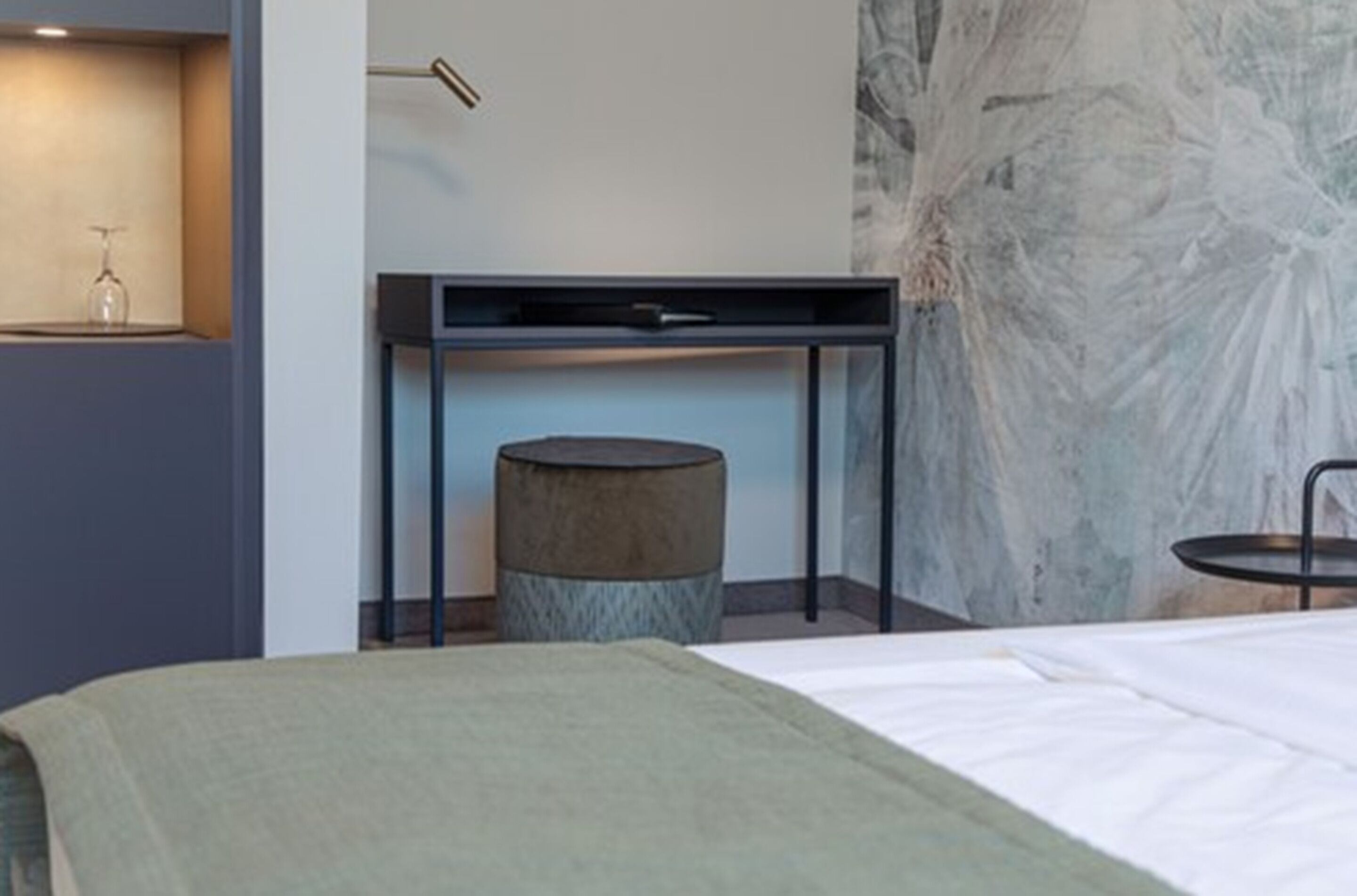 Una habitación de hotel moderna con una cama, una mesita redonda pequeña, un escritorio con una silla y un estante en la pared con un objeto de vidrio.