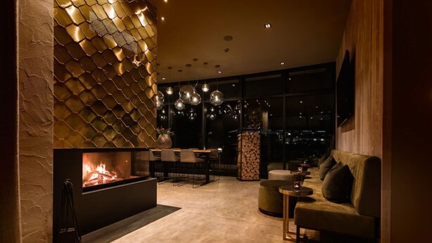 A modern living room with a fireplace, a sofa, a dining area, and a large window offering a city view. - Parkhotel Schmid (Adelsried)