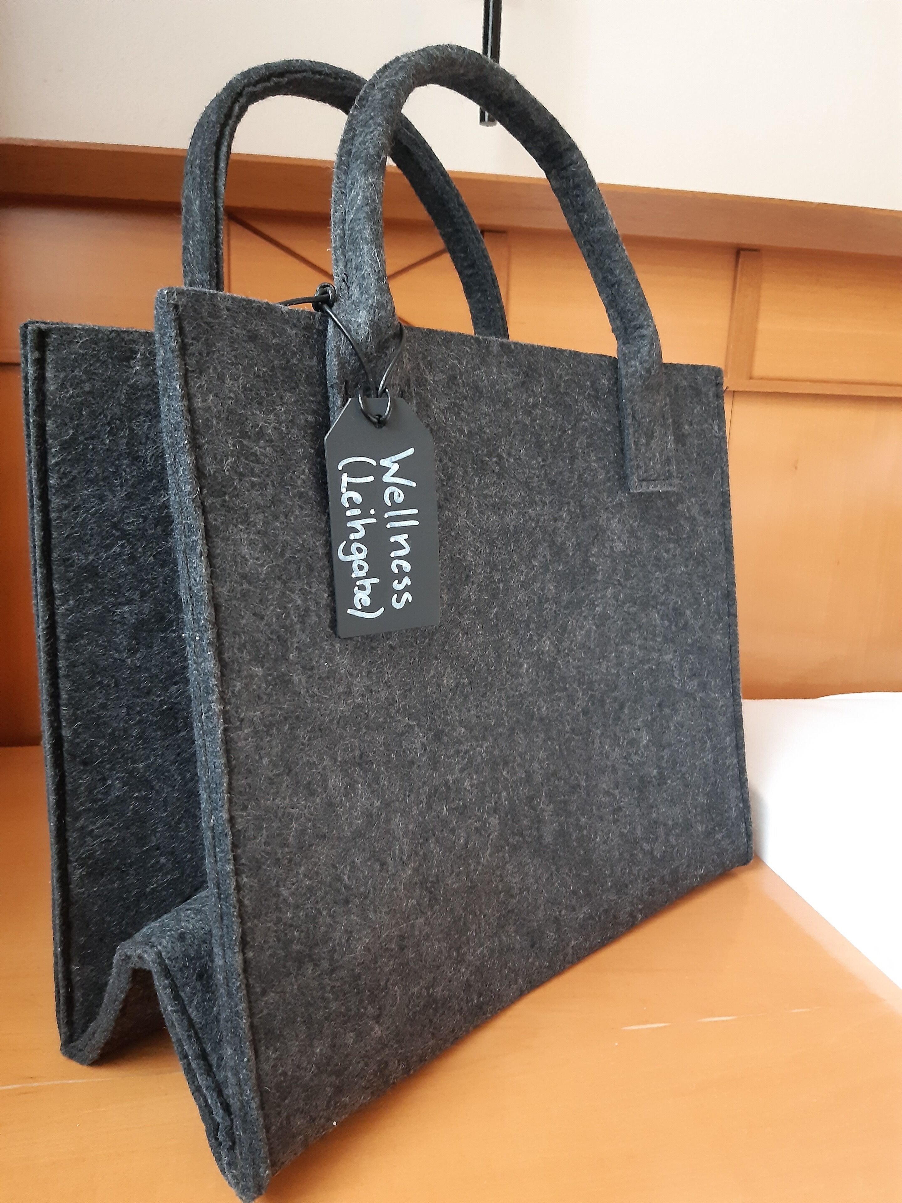 A dark gray felt tote bag with a tag labeled "Wellness (Leihgabe)" on a wooden surface.