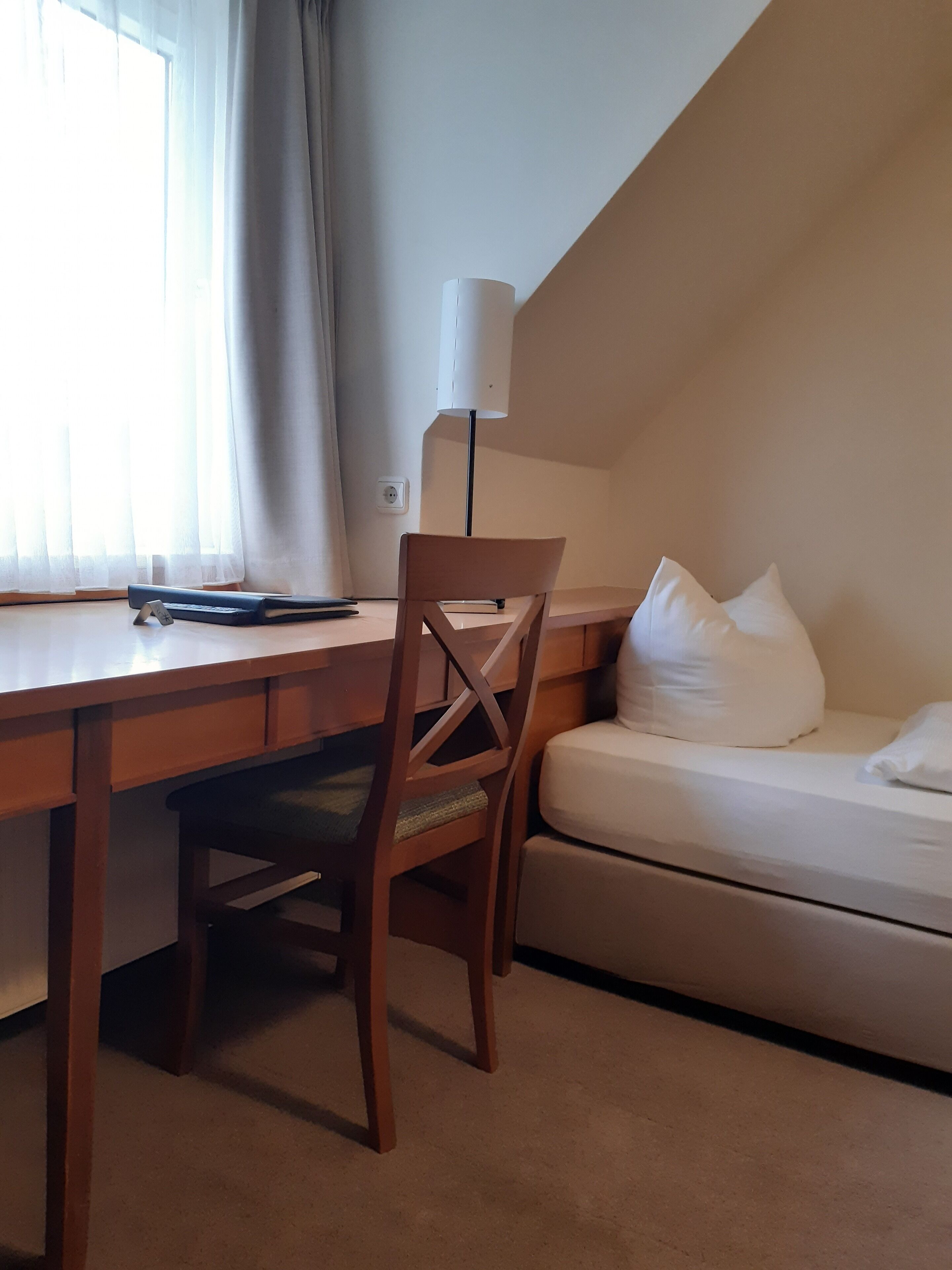 Una habitación con un escritorio de madera, una silla, una lámpara, una cama con almohadas blancas y una ventana con cortinas.