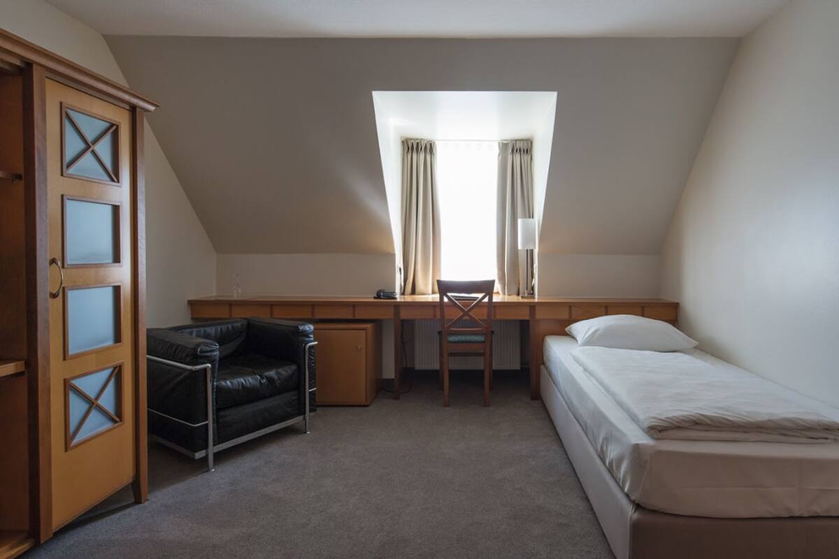 Quarto pequeno e bem iluminado, com escrivaninha, cadeira, cama e guarda-roupa.