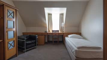 A small, well-lit room with a desk, a chair, a bed, and a wardrobe.