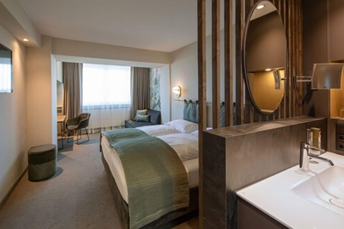 Quarto de hotel com cama, escrivaninha, cadeira, espelho grande e banheiro com pia.