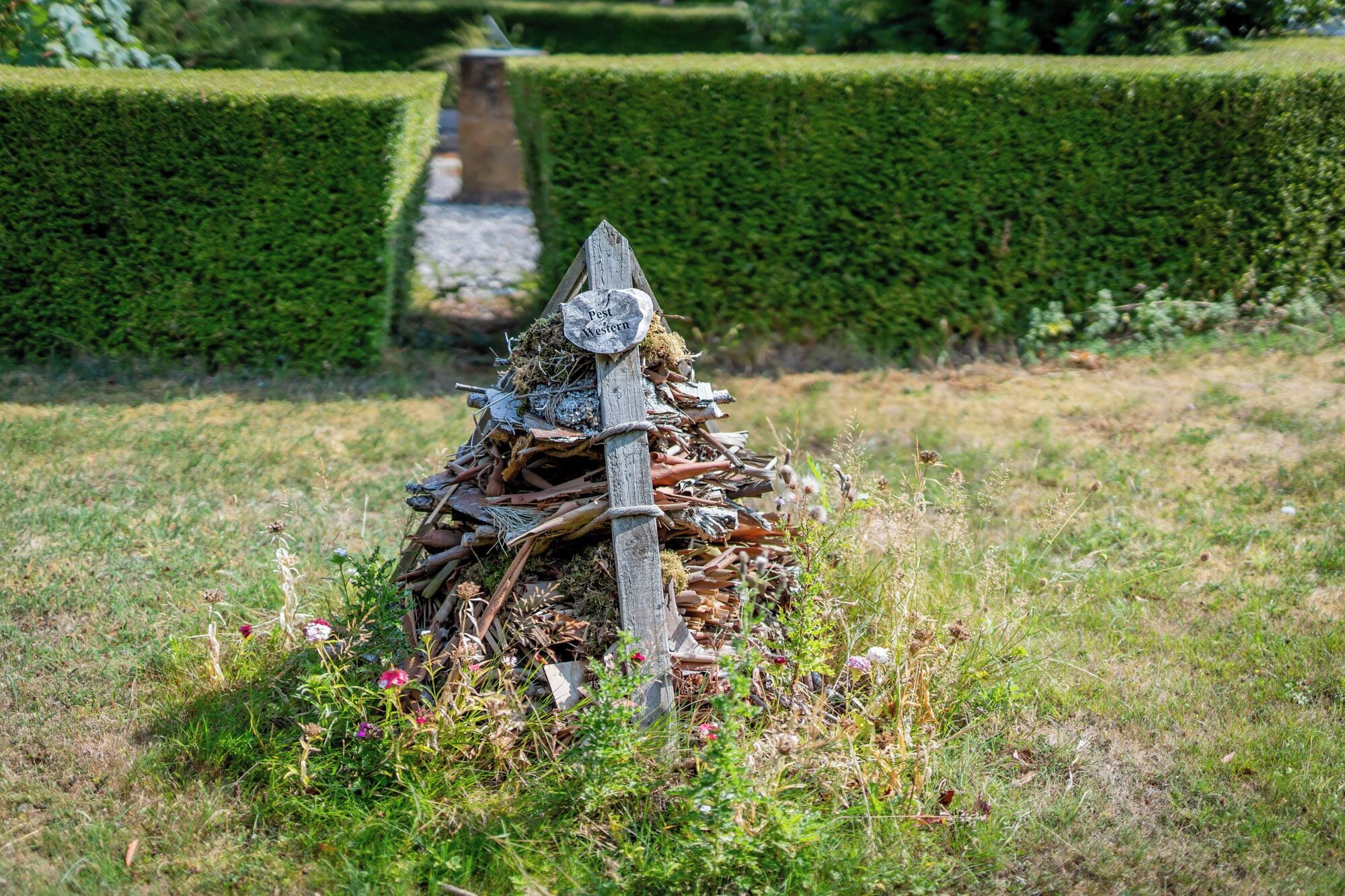 A rustic wooden structure with a sign indicating 'Pest Western' amidst a garden with neatly trimmed hedges and a gravel path.