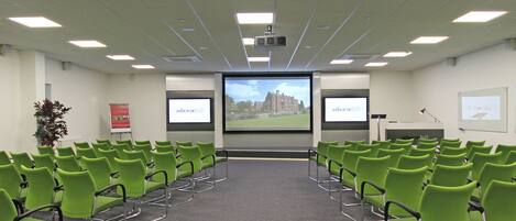 A conference room with green chairs arranged in rows, a projector screen, and a whiteboard.