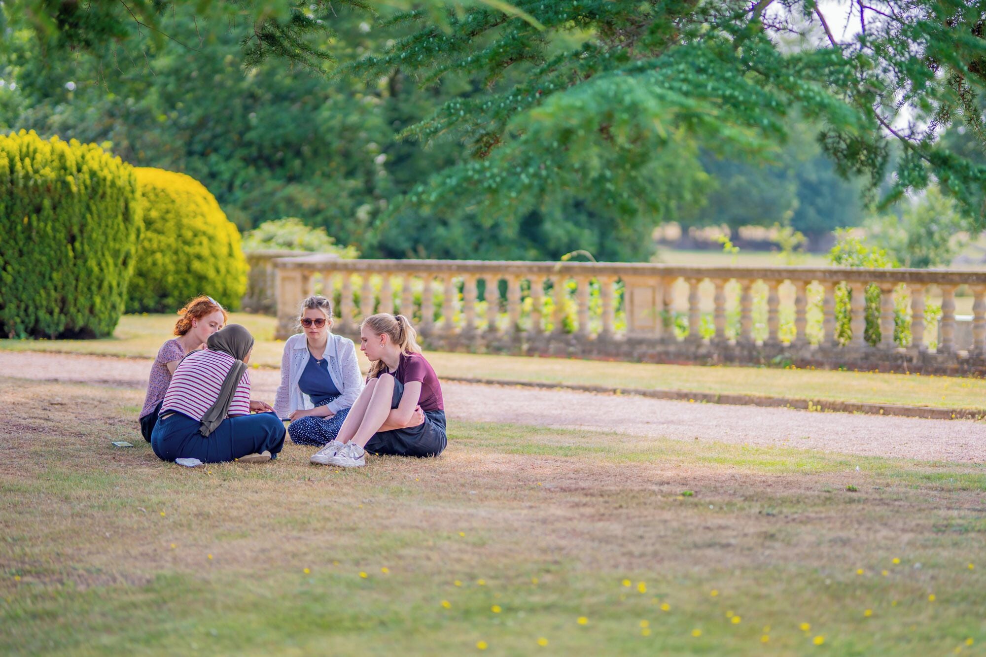 A group of people sitting on the grass in a park with a stone balustrade in the background.