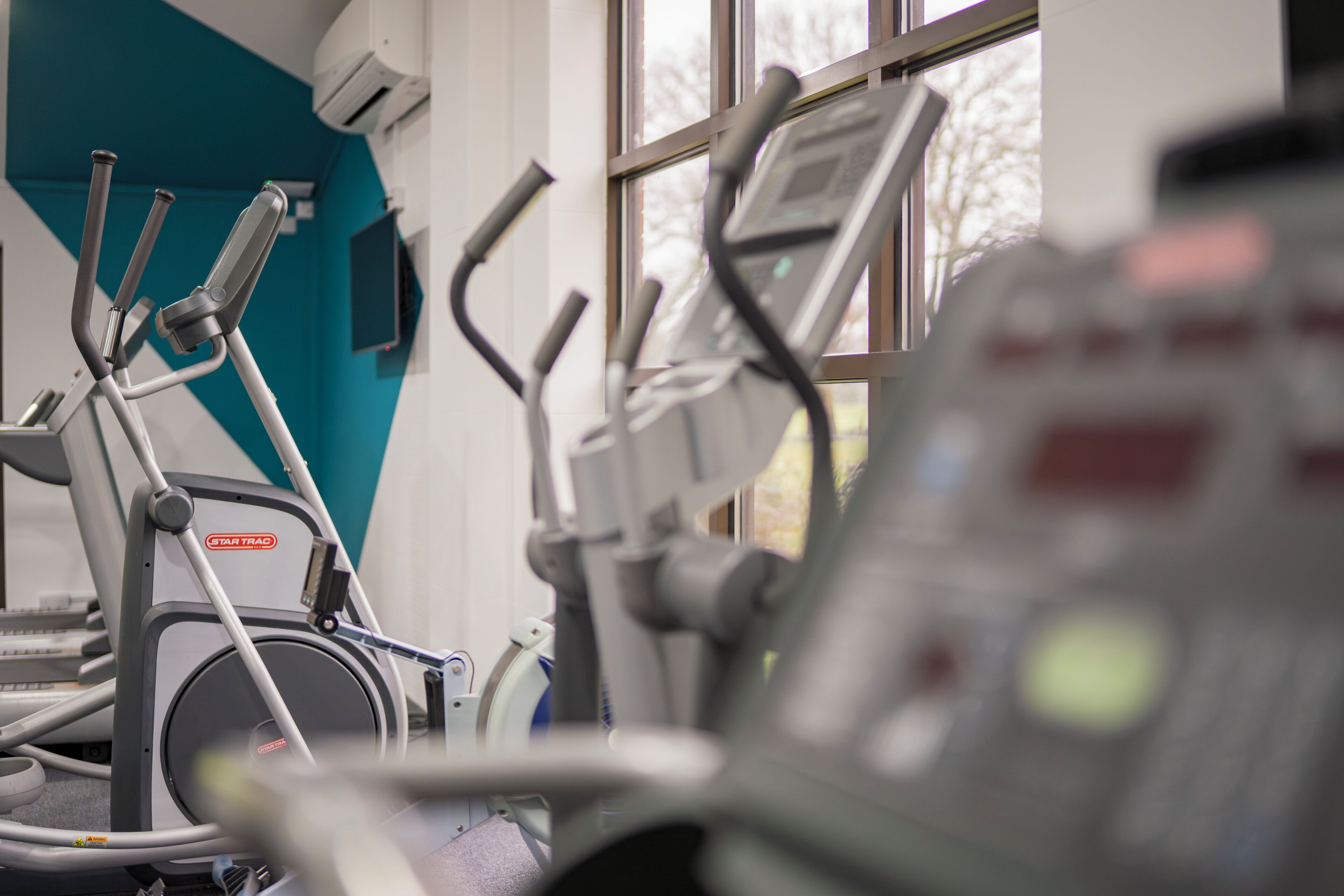 An exercise machine with a digital display, a wall-mounted air conditioning unit, and a window with a view of trees.