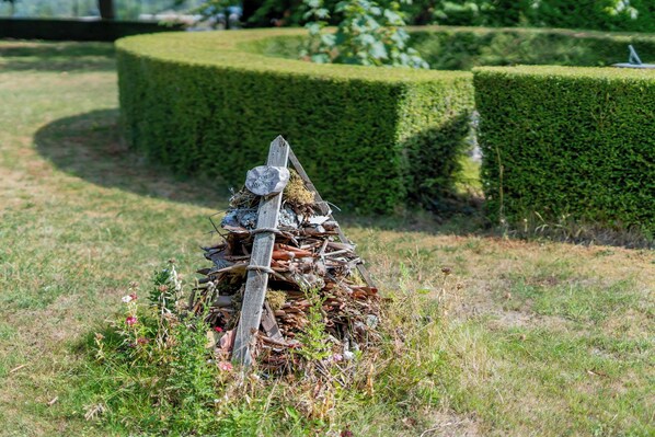 A rustic wooden sign with a bird perched on top, surrounded by a pile of twigs and branches, set against a backdrop of neatly trimmed hedges and a grassy area.