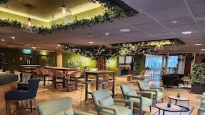 A modern lounge area with wooden flooring, green upholstered chairs, and round tables with potted plants. - Ashorne Hill Leamington Spa (Royal Leamington Spa)