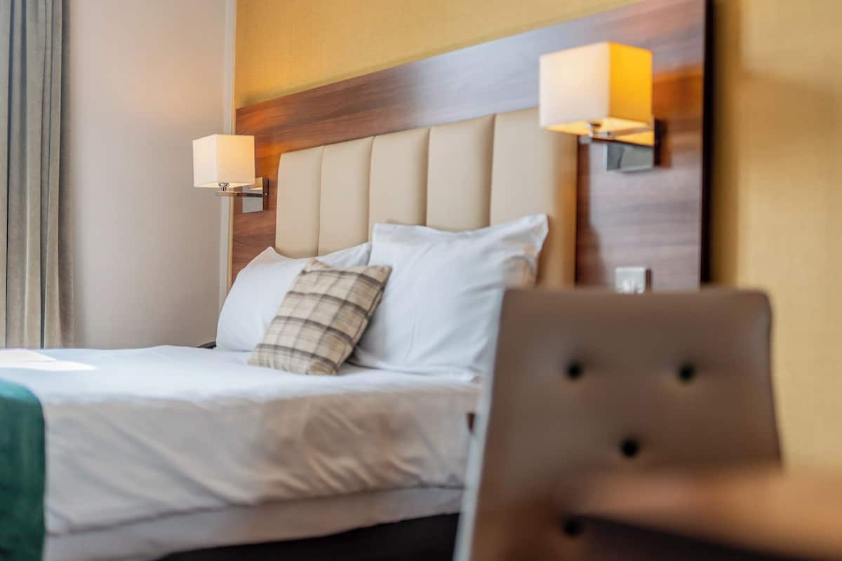 A neatly made bed with white linens and a plaid pillow, flanked by two wall-mounted lamps.