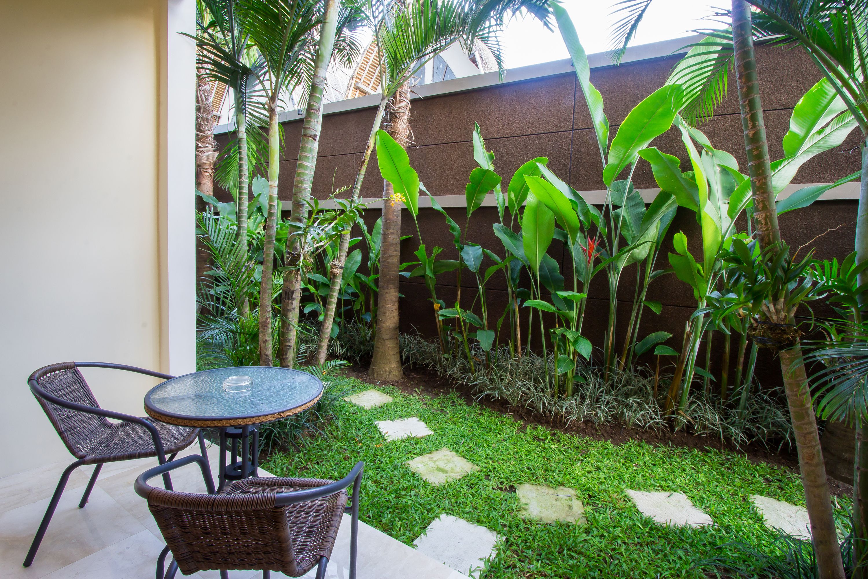 A patio with a round glass table and wicker chairs, surrounded by lush greenery and a stone pathway.