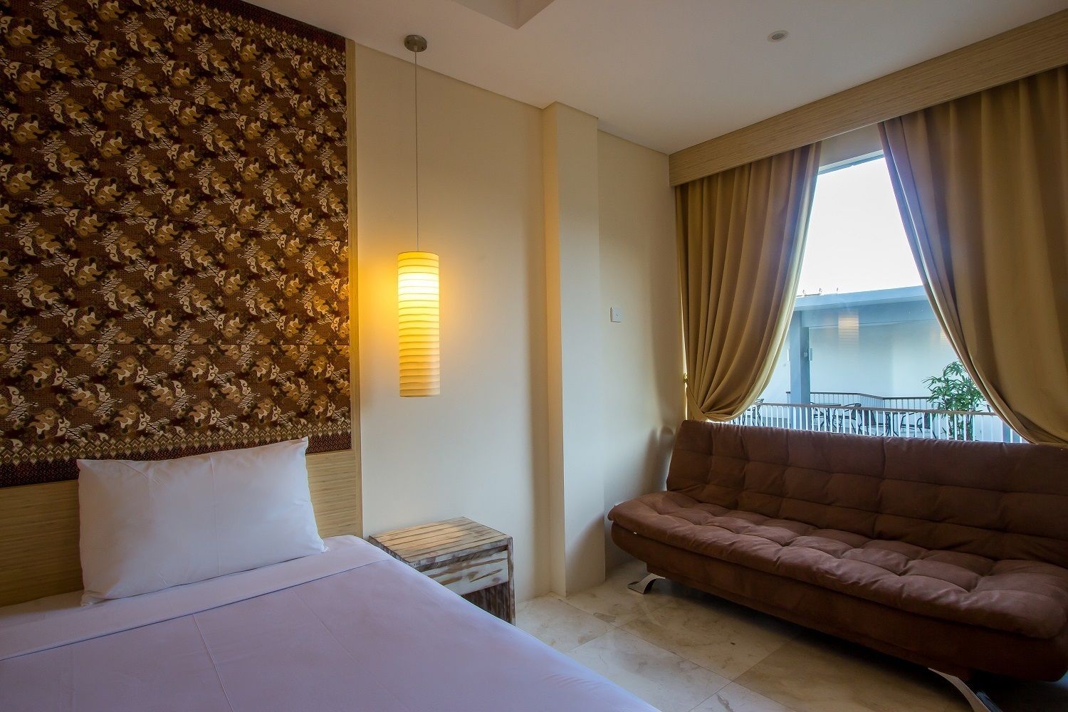 A hotel room with a bed, a sofa, a patterned headboard, a wall-mounted lamp, and a window with curtains.