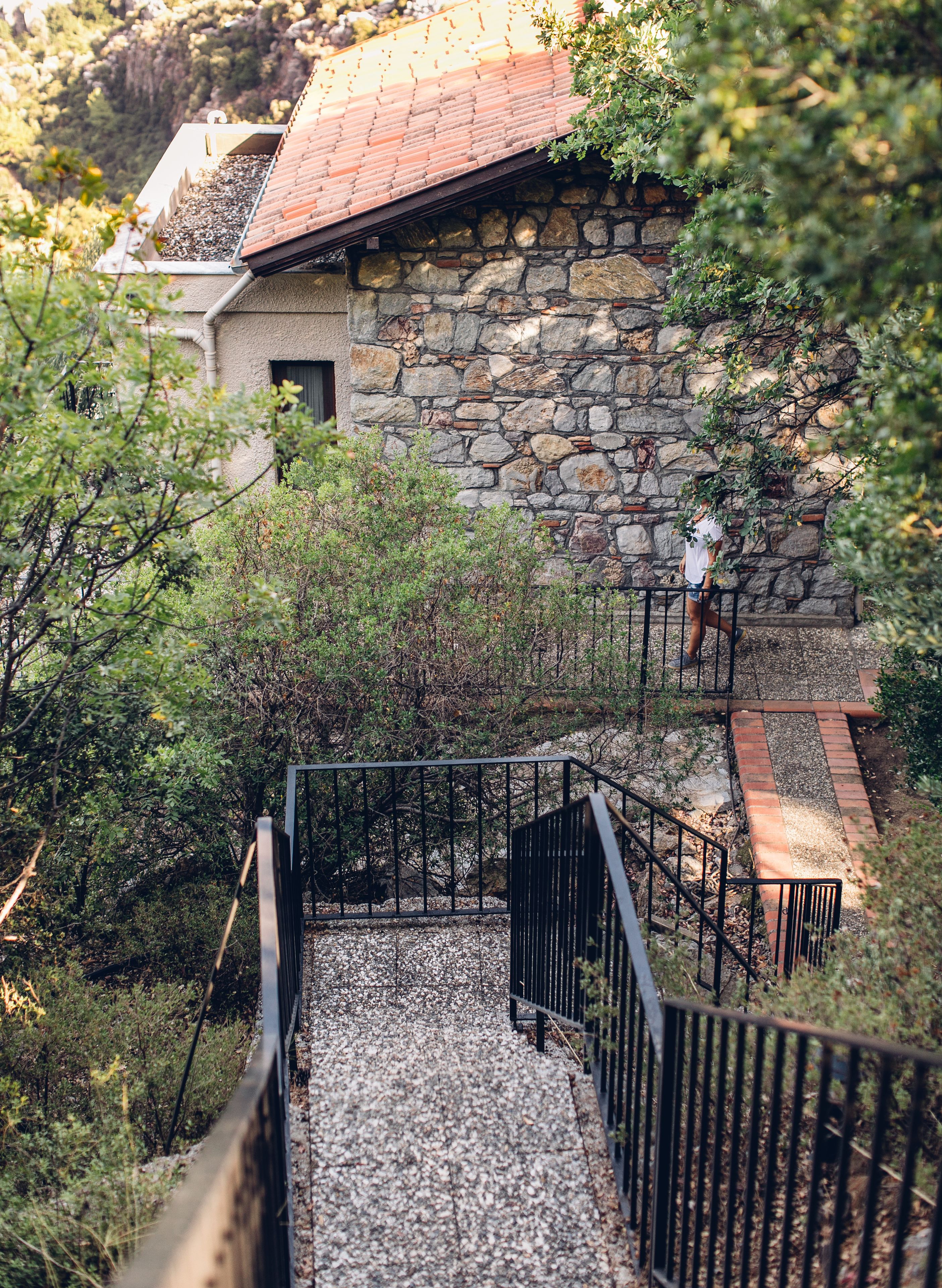 A stone building with a red-tiled roof, surrounded by greenery and a gravel pathway leading to a black metal staircase.