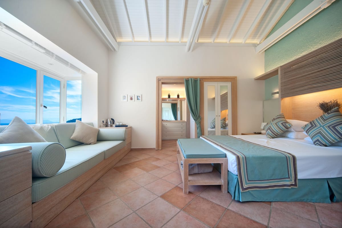 Une chambre avec une grande fenêtre, un canapé, un lit recouvert d’une couverture verte et une tête de lit en bois.