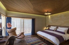 Une chambre d’hôtel moderne avec un plafond en bois, un grand lit avec une literie blanche et violette, un bureau avec un ordinateur portable et une chaise.