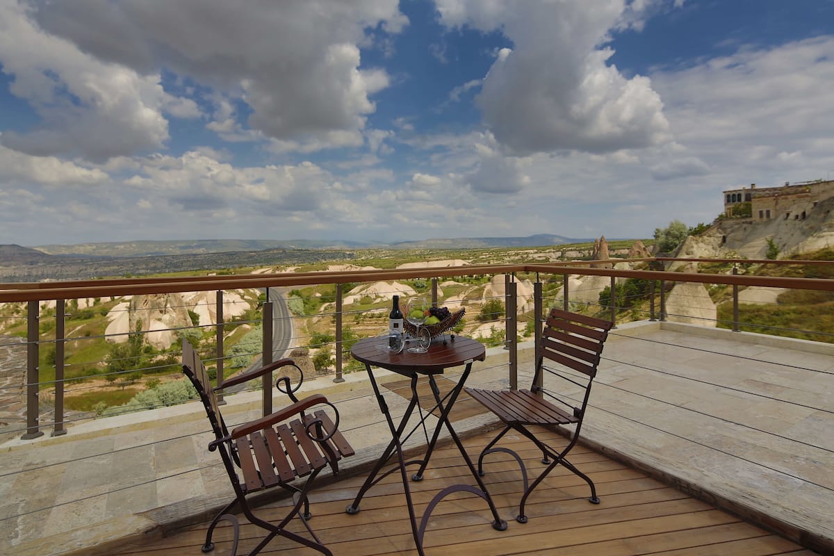 Een balkon met twee houten stoelen en een kleine ronde tafel, met uitzicht op een schilderachtig landschap met bergen en een gebouw op een heuvel.