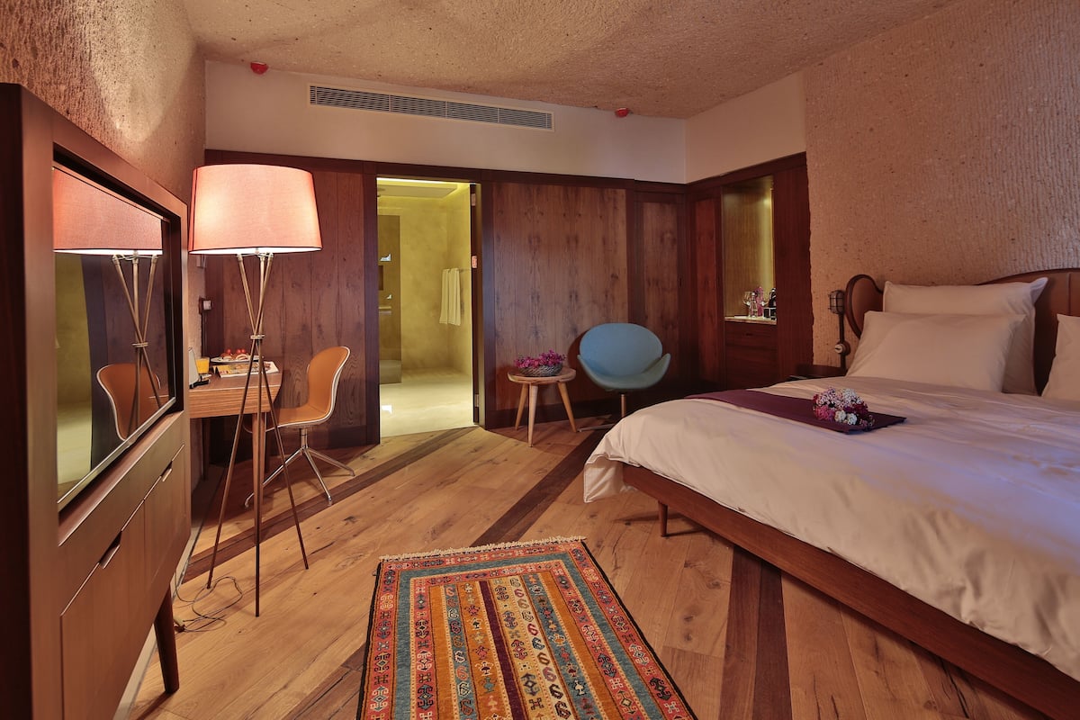 Een hotelkamer met een houten vloer, een bed met witte lakens, een bureau met een lamp, een stoel en een kleine tafel met een bloevas.