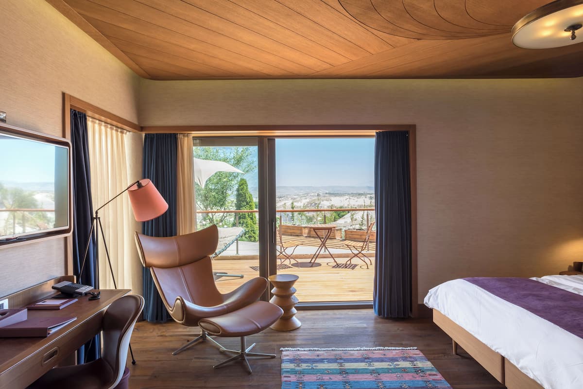 Een moderne hotelkamer met een groot bed, een bureau met een computer, een stoel en uitzicht op het omliggende landschap door een schuifdeur.