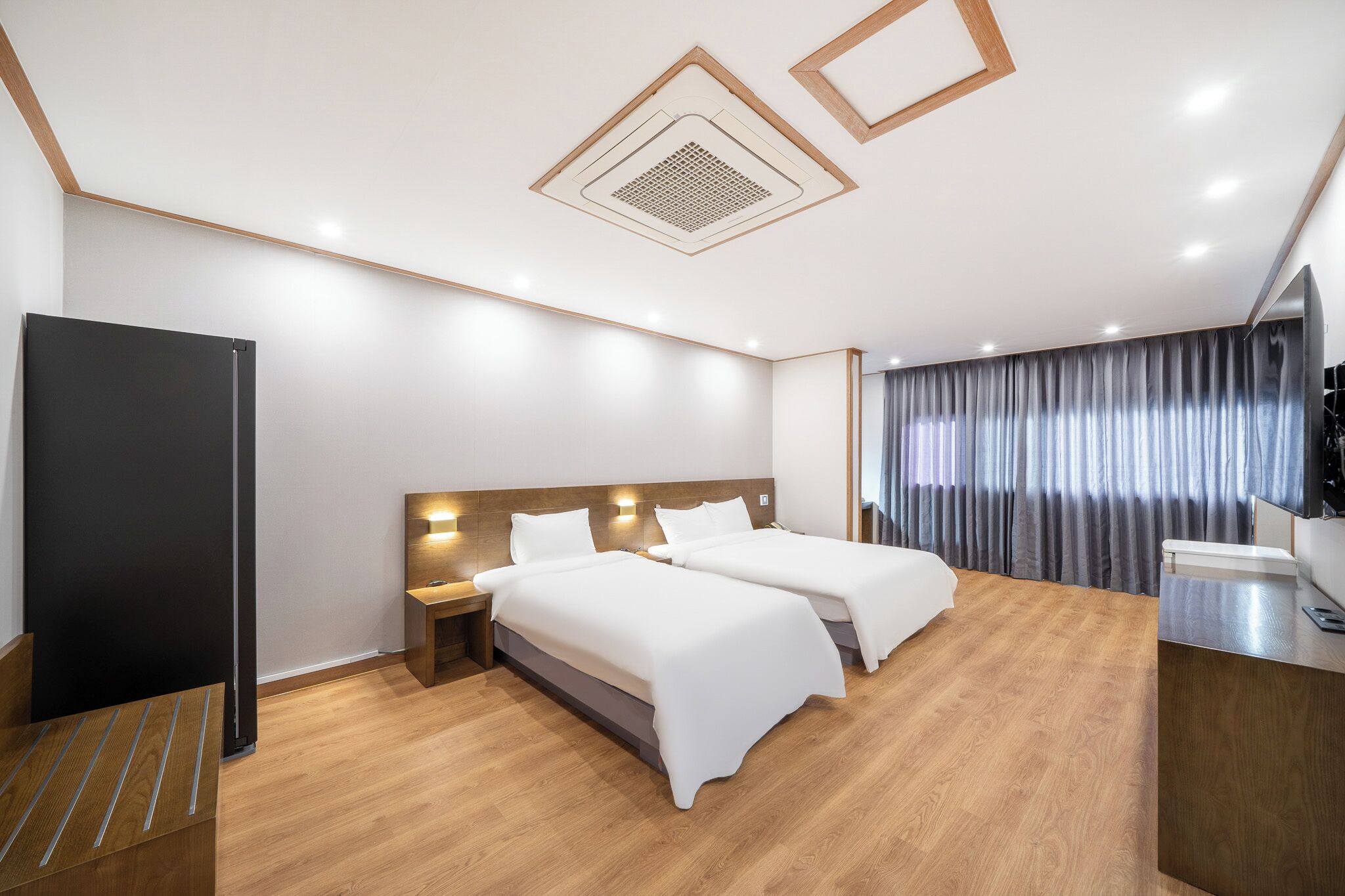 A modern hotel room with two beds, wooden flooring, a flat-screen TV, and a large window with curtains.