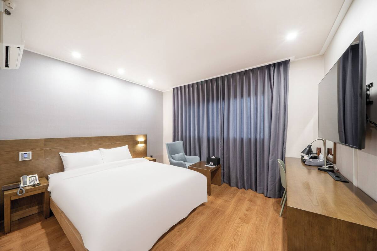 Habitación de hotel con una cama grande, un escritorio con una silla, una mesita de noche con lámpara, un teléfono y un televisor.