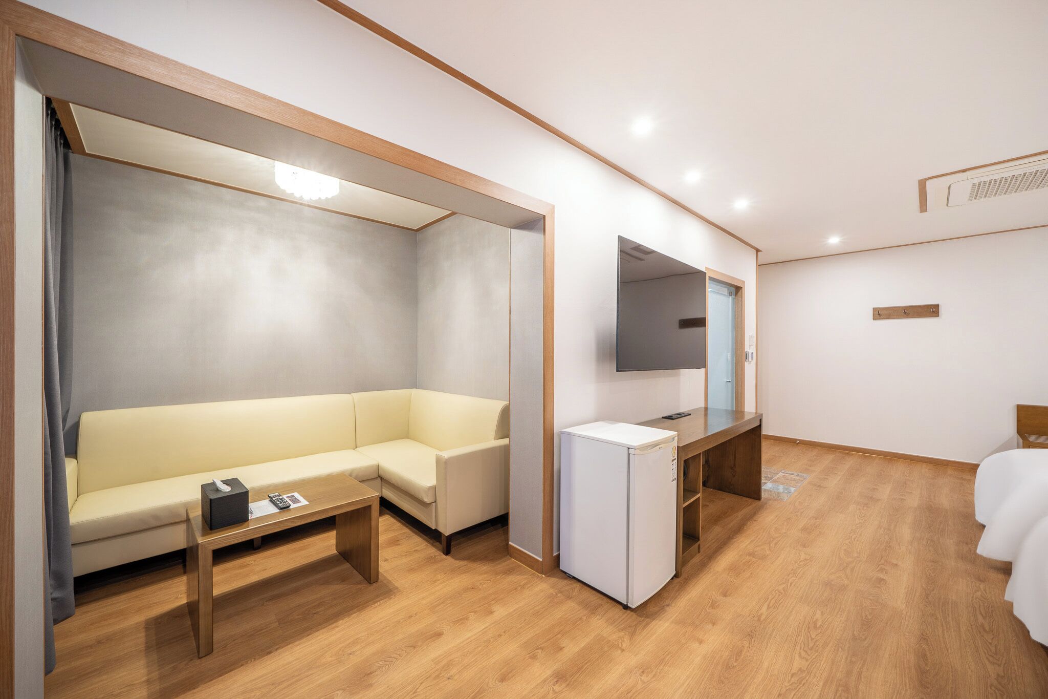 A modern hotel room with a wooden floor, a white sofa, a small desk with a mirror, and a bed.