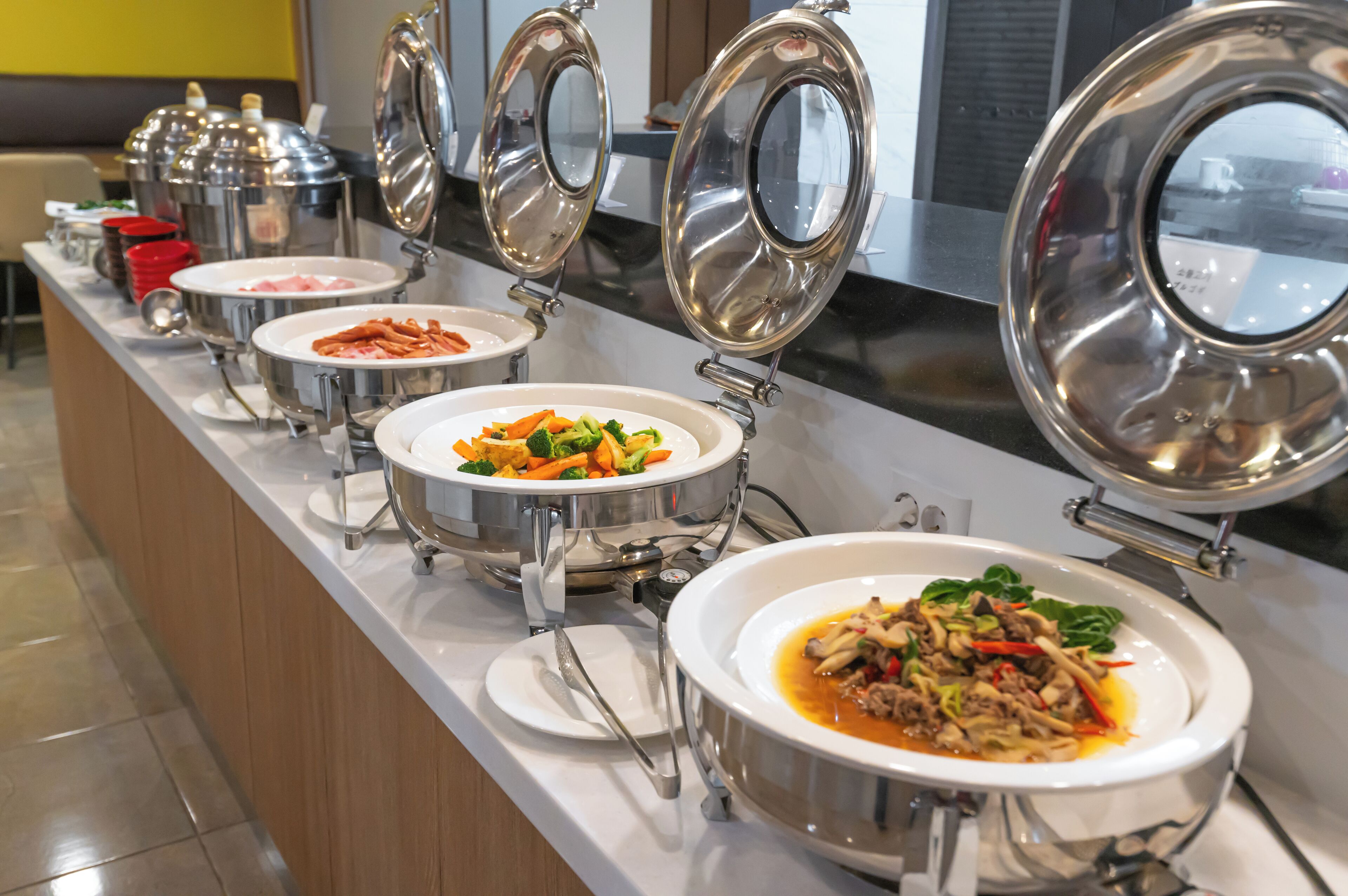 A buffet line with various dishes in stainless steel chafing dishes, including a noodle dish, a vegetable stir-fry, and a meat dish.