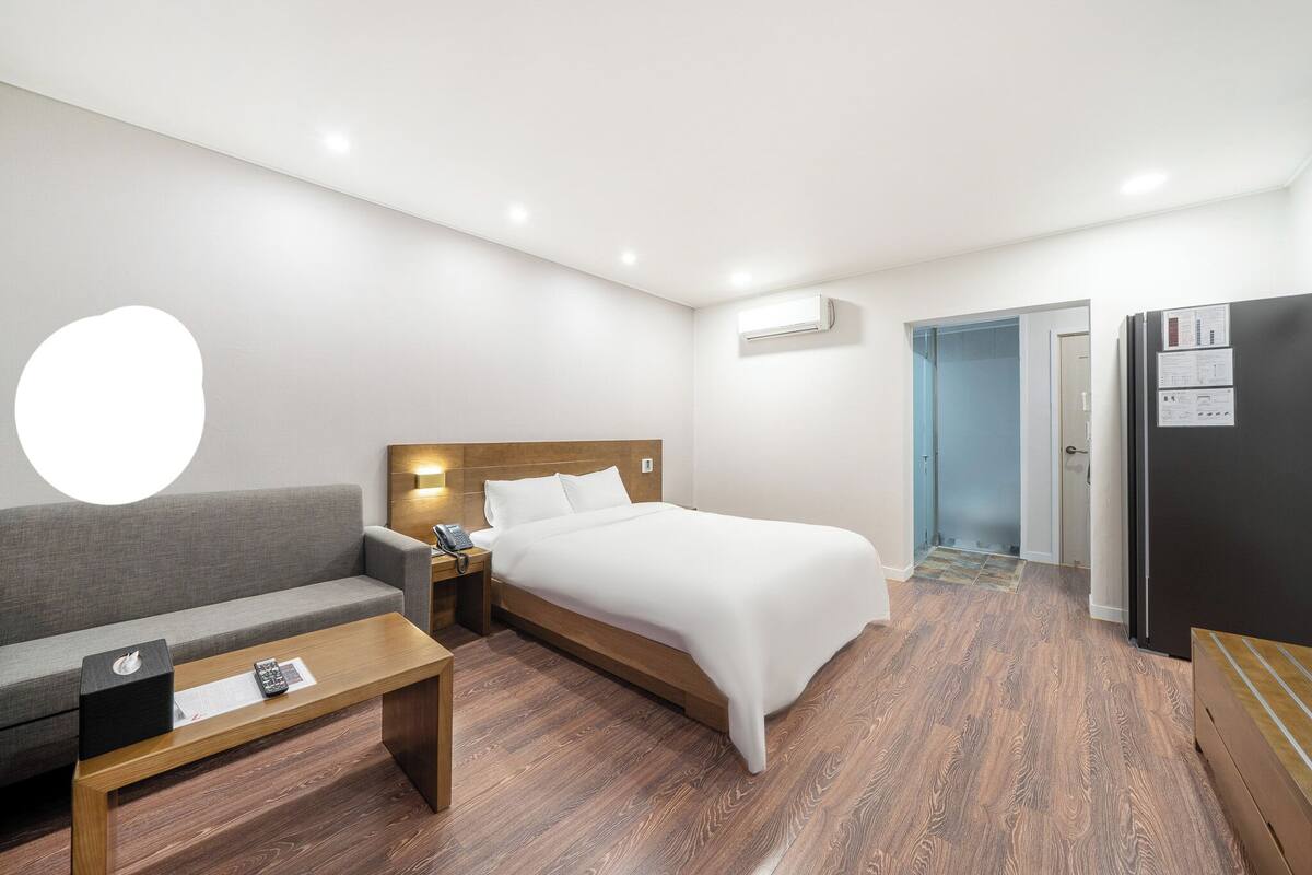 Habitación de hotel con cama, sofá, mesita de centro de madera, mesita de noche, equipo de aire acondicionado, refrigerador y puerta que da a otra habitación.