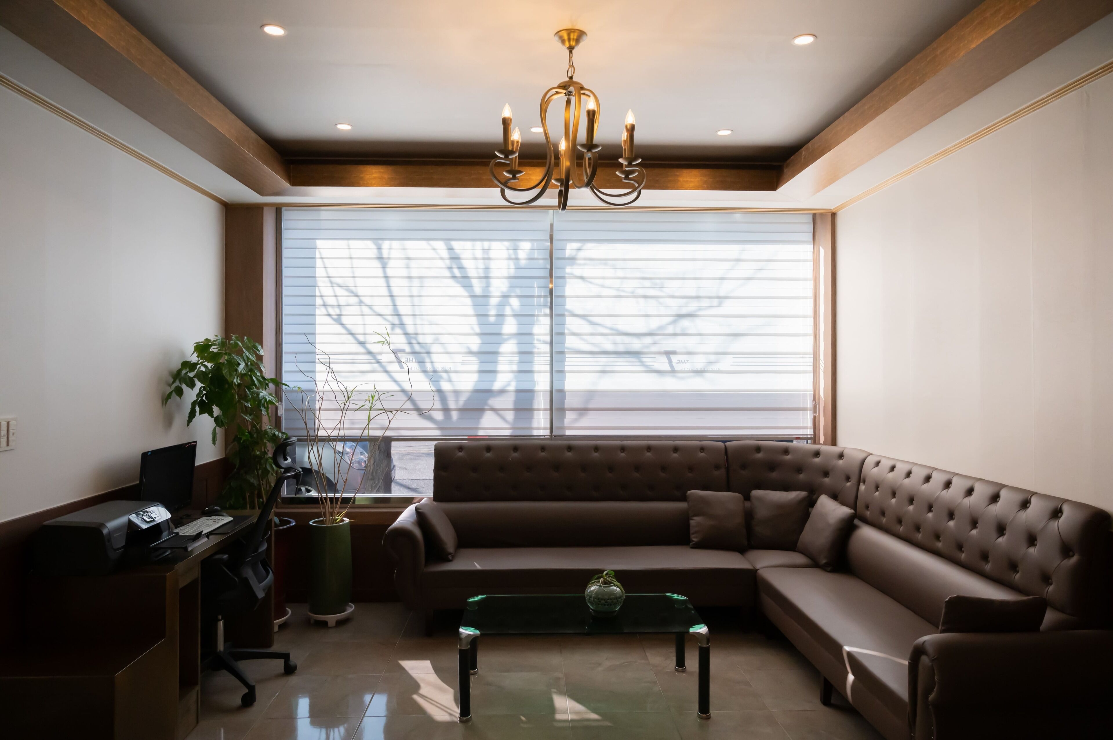 A modern living room with a brown leather sofa, a glass coffee table, a desk with a computer, and a large window with blinds.