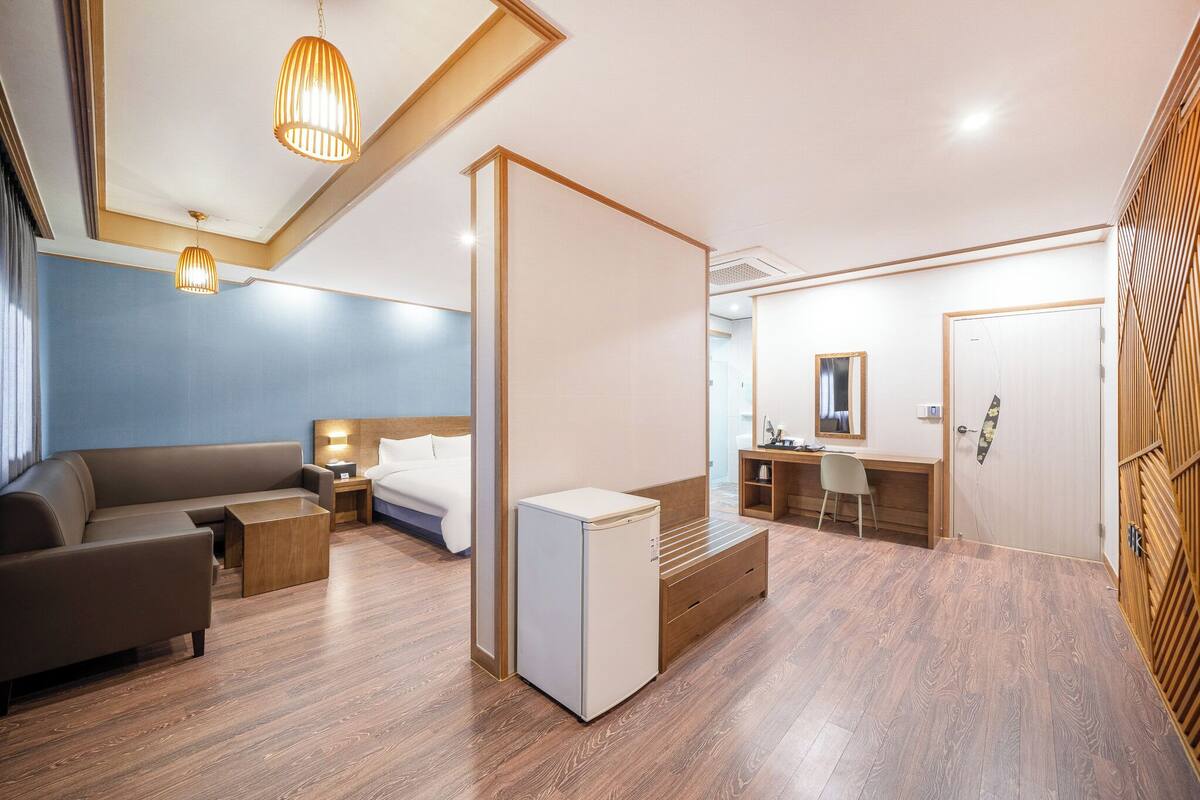 Una habitación de hotel moderna con sofá modular, un refrigerador blanco pequeño, un escritorio con silla y un espejo grande en la pared.