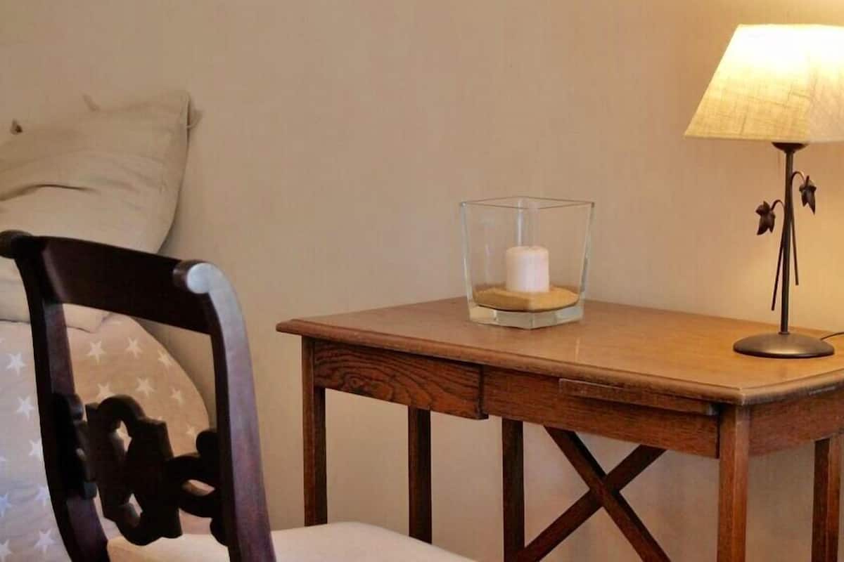Une table de chevet en bois, sur laquelle se trouvent un porte-bougie en verre, une lampe et une chaise avec un coussin.