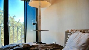 Une chambre avec une grande fenêtre, une table de chevet sur laquelle se trouve un verre à vin, et un livre posé sur le lit.