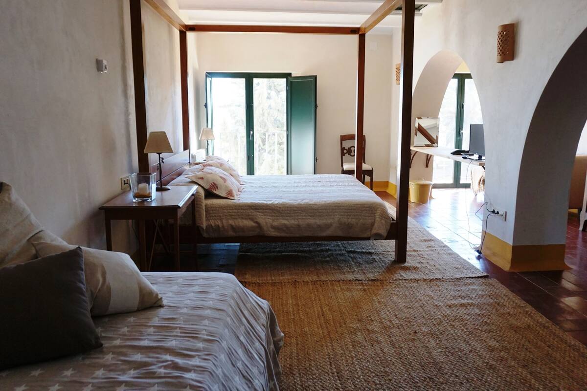 Une chambre avec deux lits, un bureau en bois, une lampe et une vue sur l’extérieur par une porte ouverte.